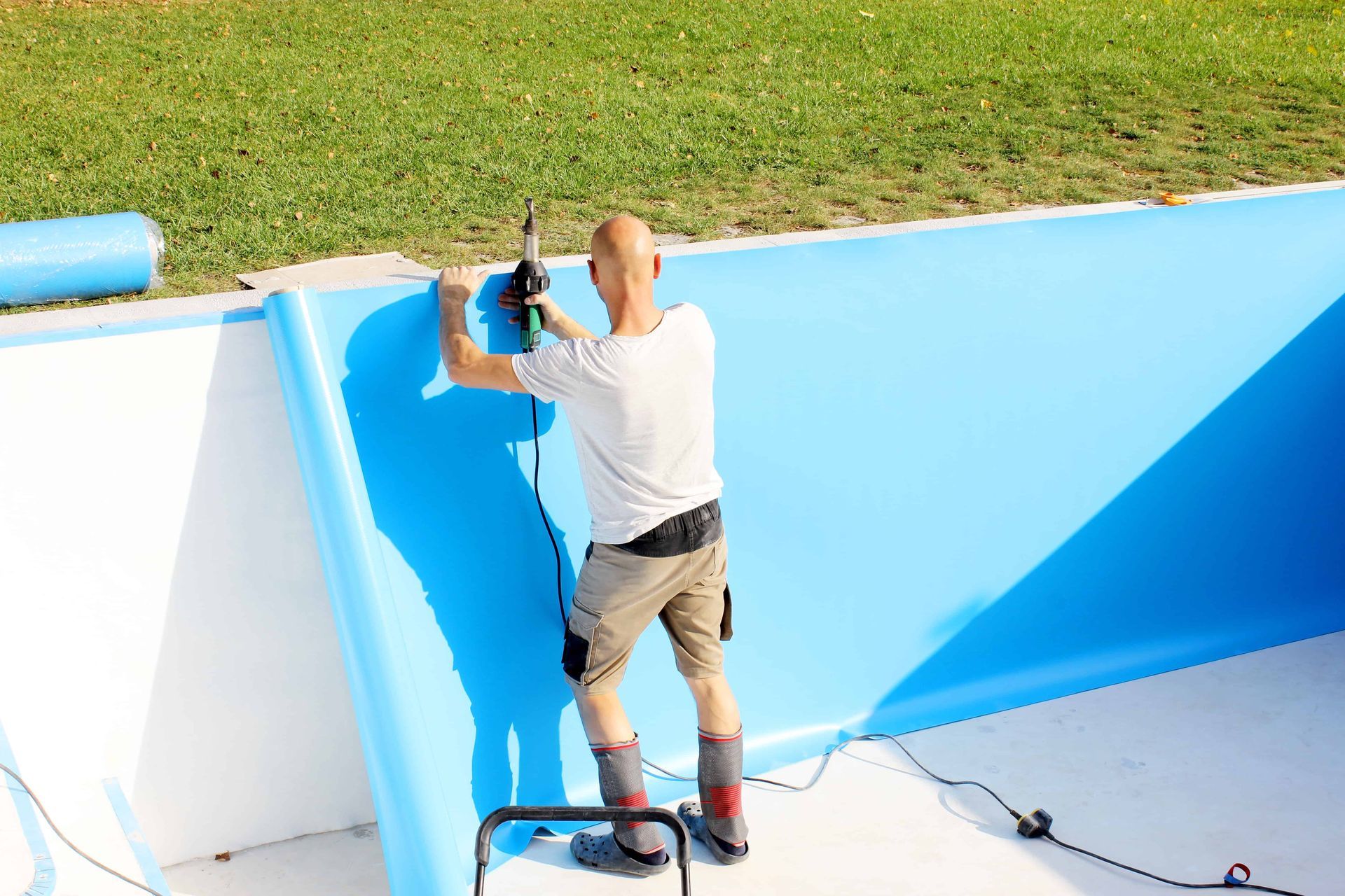 Person welding blue pool liner to white wall of an empty pool. Green grass in background.