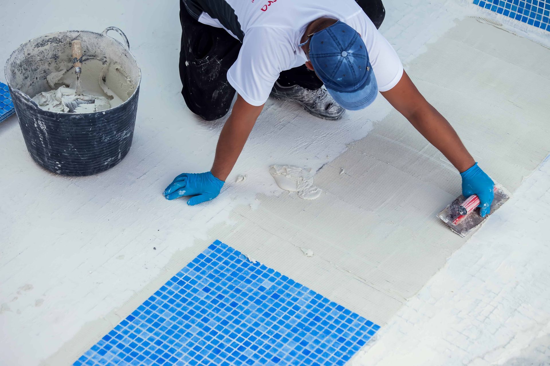 Person tiling a surface, applying adhesive with a trowel, blue mosaic tiles, blue gloves.
