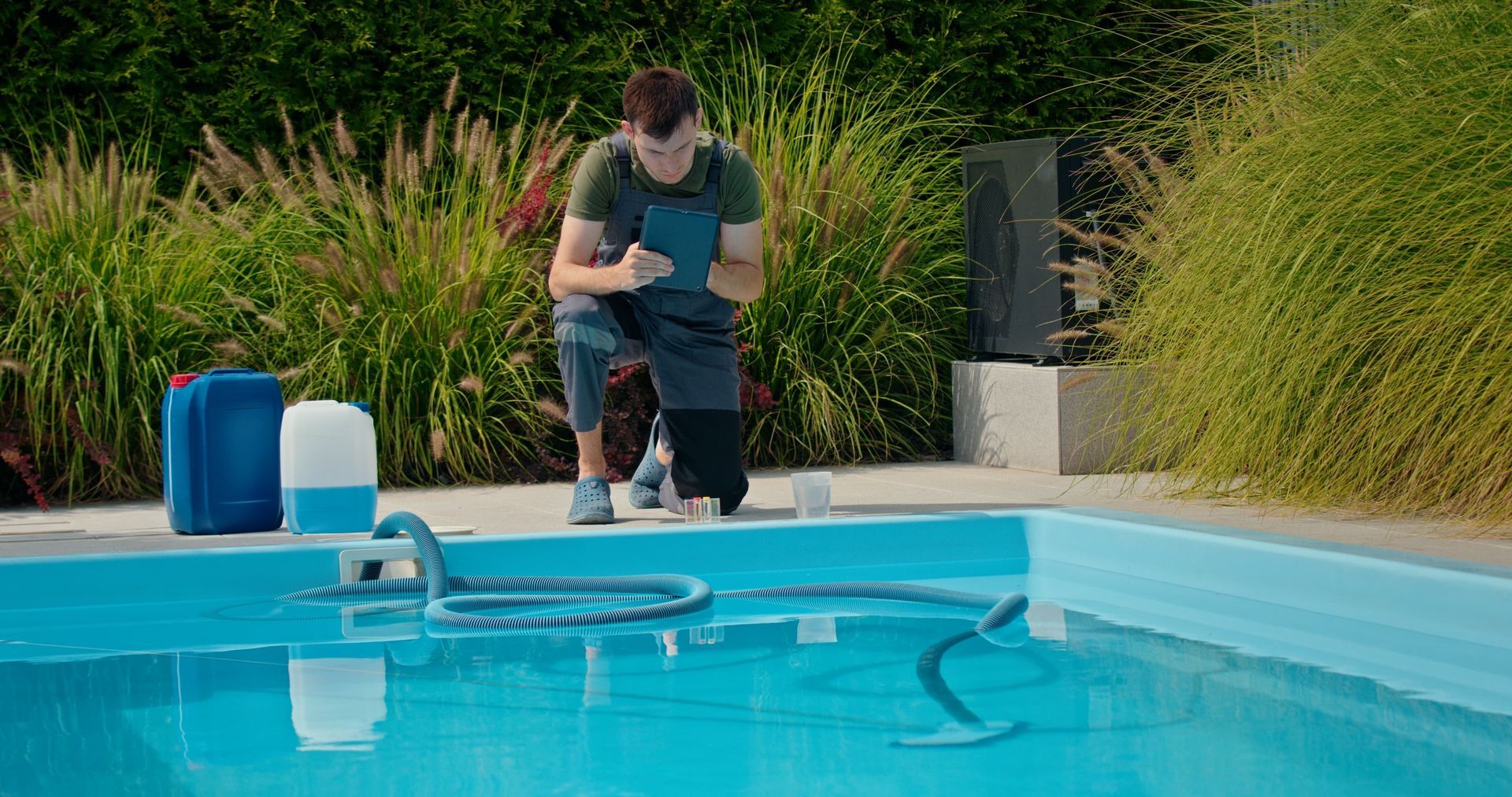 Person checks pool water quality by a pool, with cleaning equipment and chemical containers nearby.