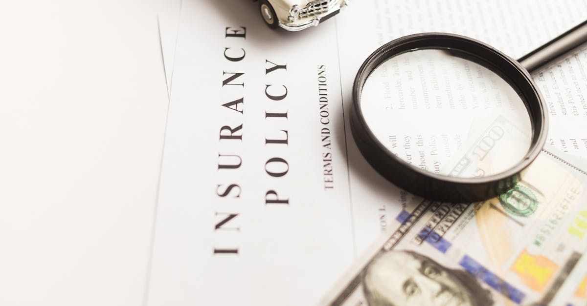 A magnifying glass rests on an insurance policy document next to a small toy car and a US one-hundred-dollar bill.