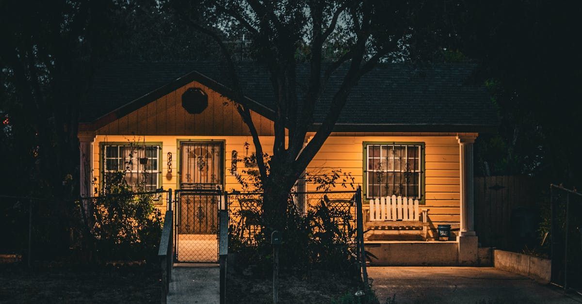 A small, yellow, single-story house at night with warm lights glowing from inside, windows, and a front porch.
