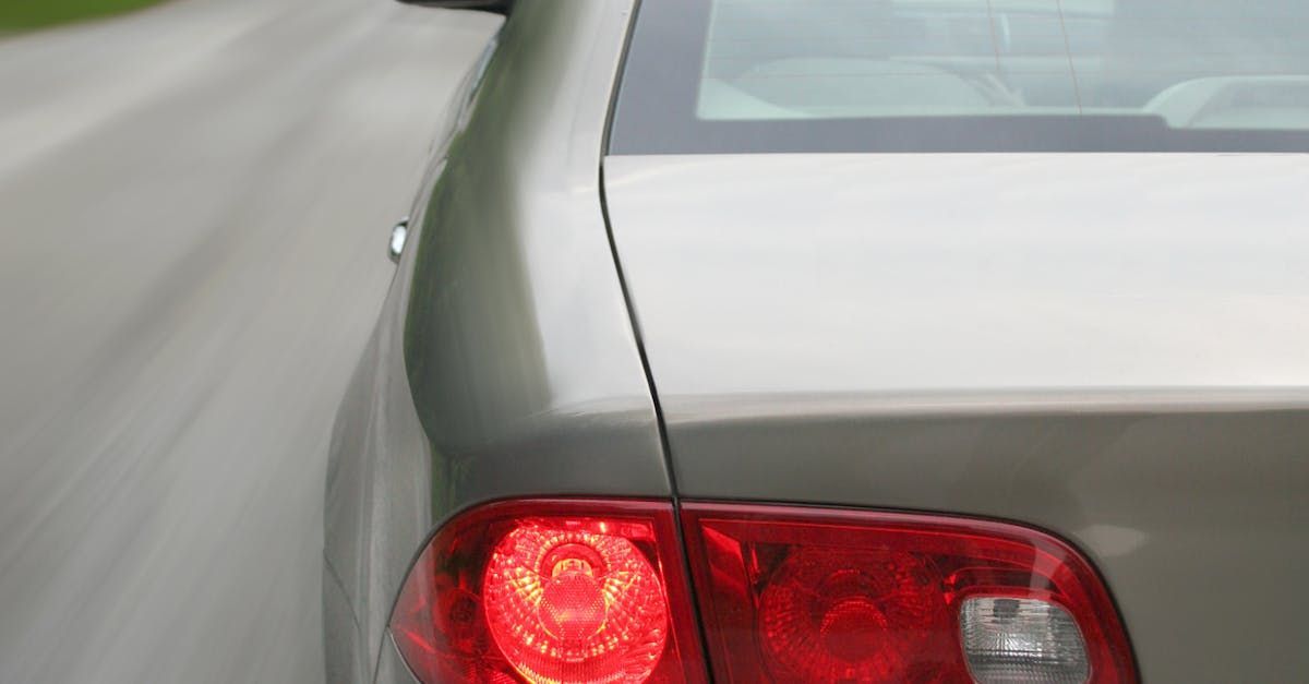 Rear view of a light-colored sedan moving on a road with its red taillight illuminated.