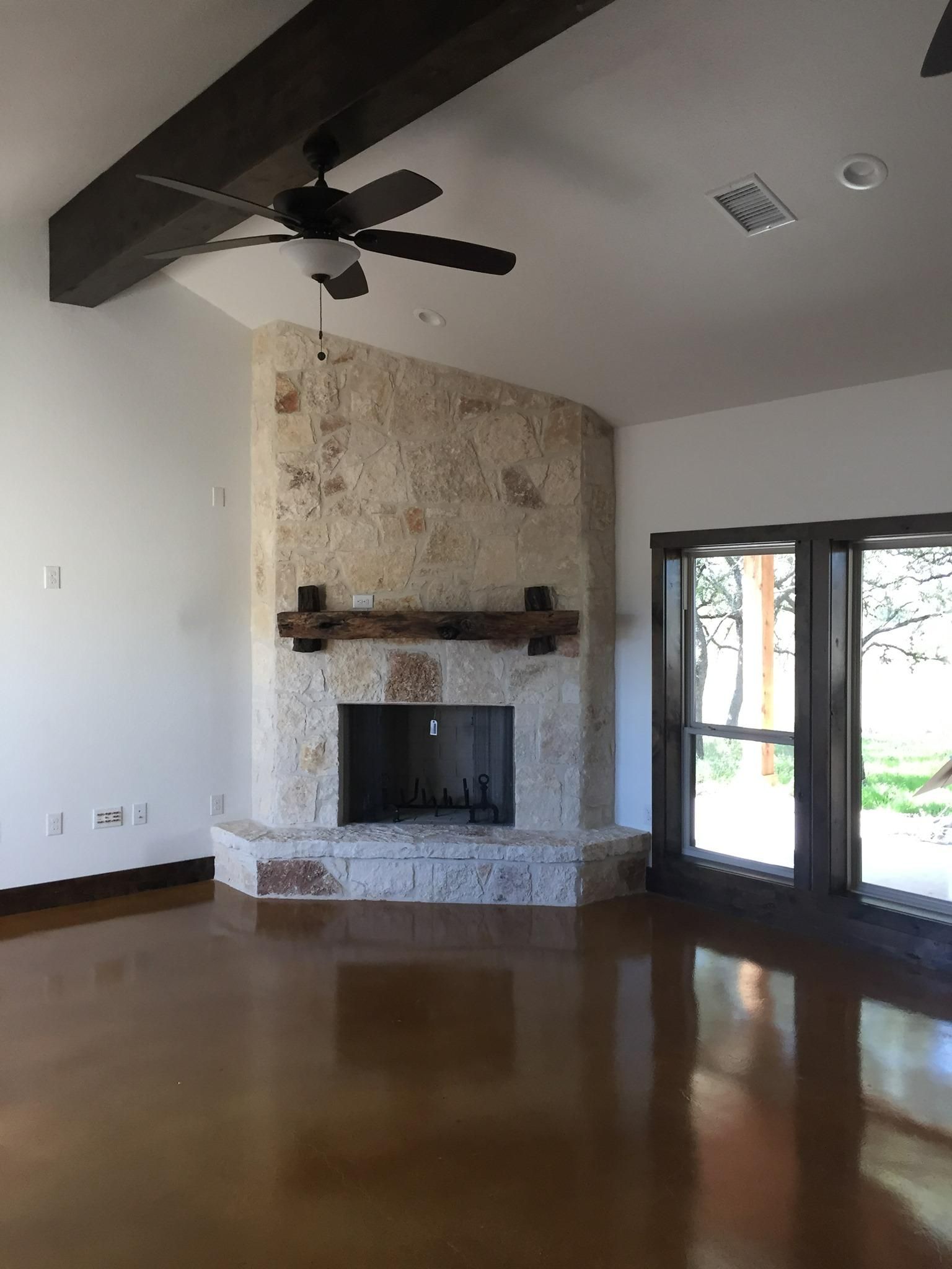 A living room with a stone fireplace, dark wood ceiling beams, and large windows. The floor is a shiny, copper-toned color.
