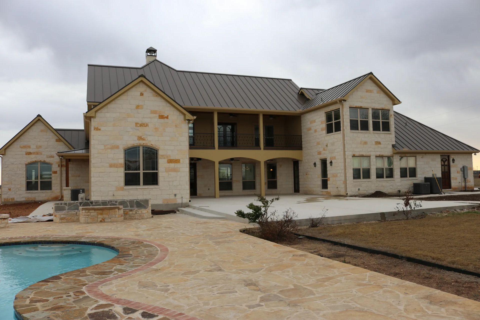 Large two-story house with stone facade, metal roof, and swimming pool in a rural setting.