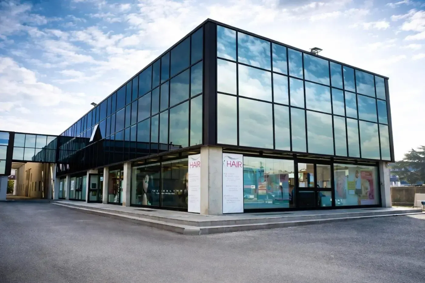 Moderno edificio a due piani con vetrate che riflettono il cielo. Il piano terra ospita negozi. Cielo nuvoloso.