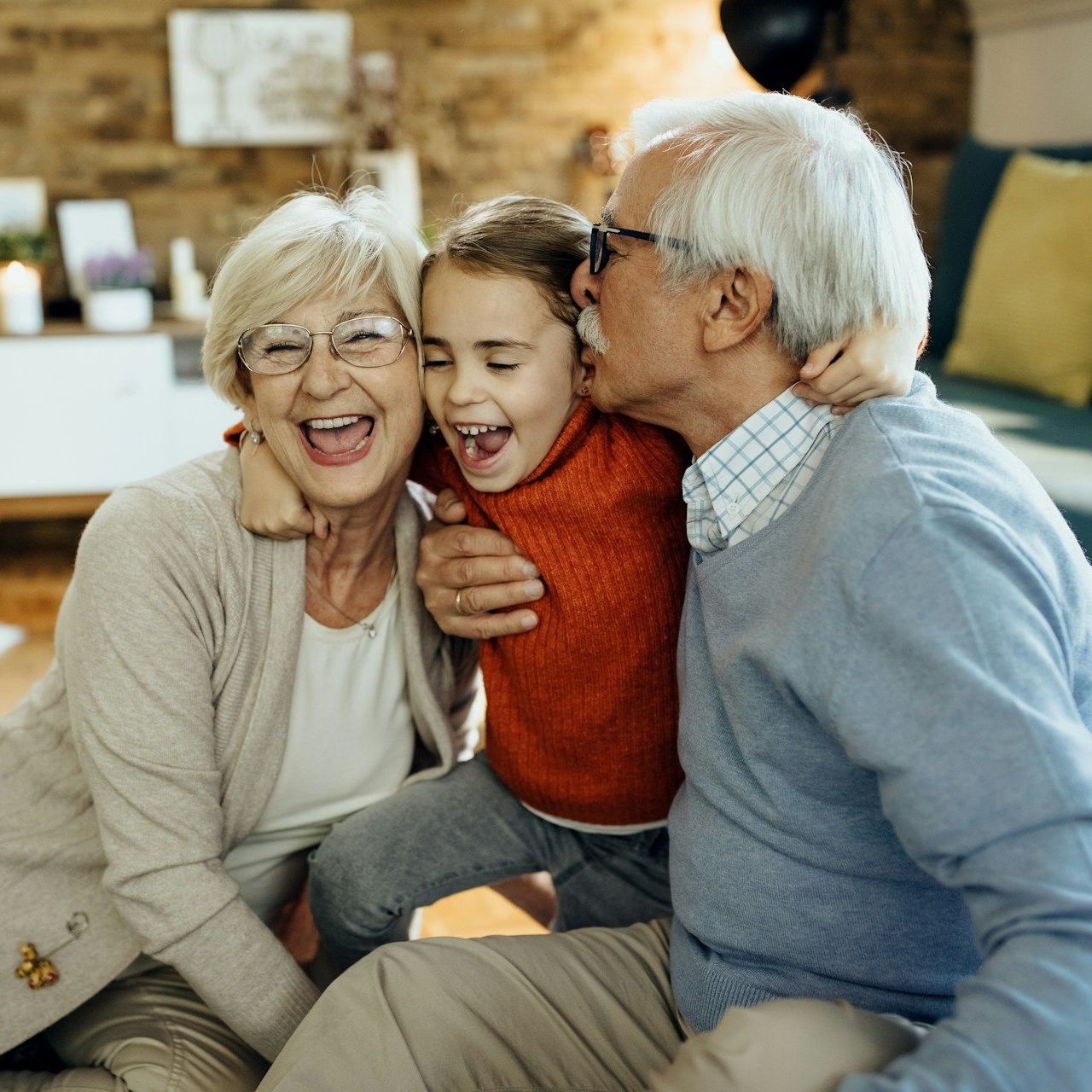 Grandparents hugging and kissing a young child indoors. They're smiling and laughing.
