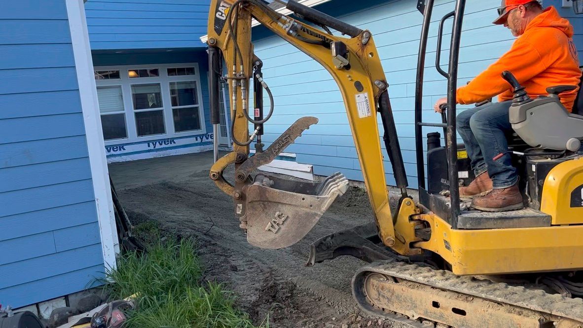 A man is driving a small excavator in front of a blue house.