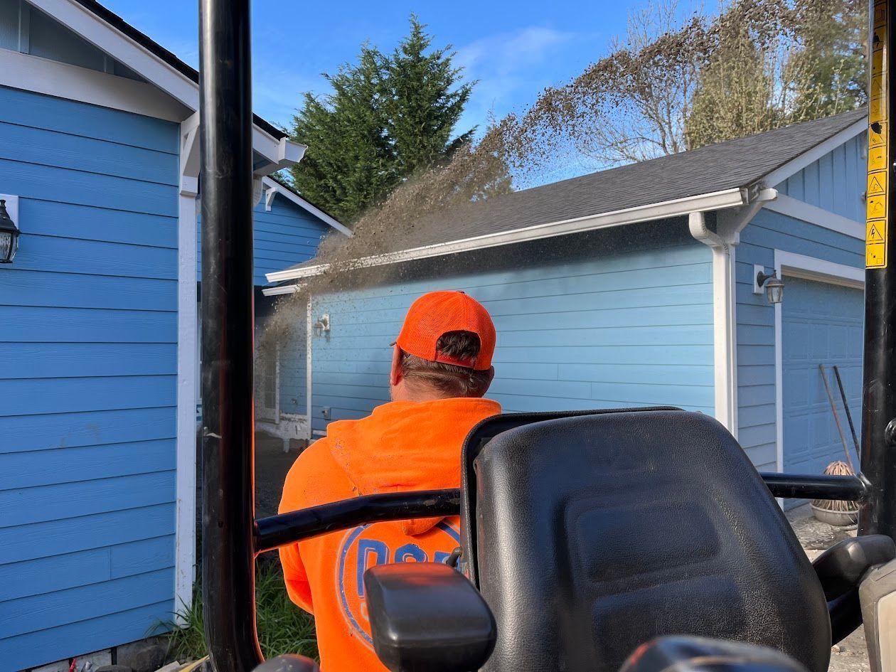 A man in an orange shirt is driving a tractor in front of a blue house.