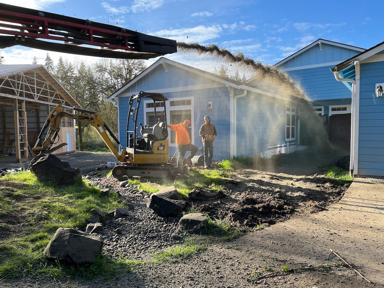 A man is standing in front of a house while an excavator is digging in the dirt.