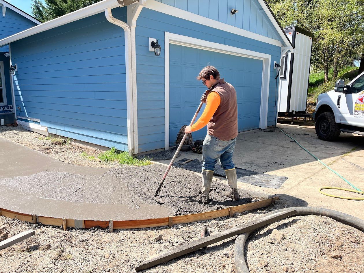 A man is raking concrete in front of a blue garage.