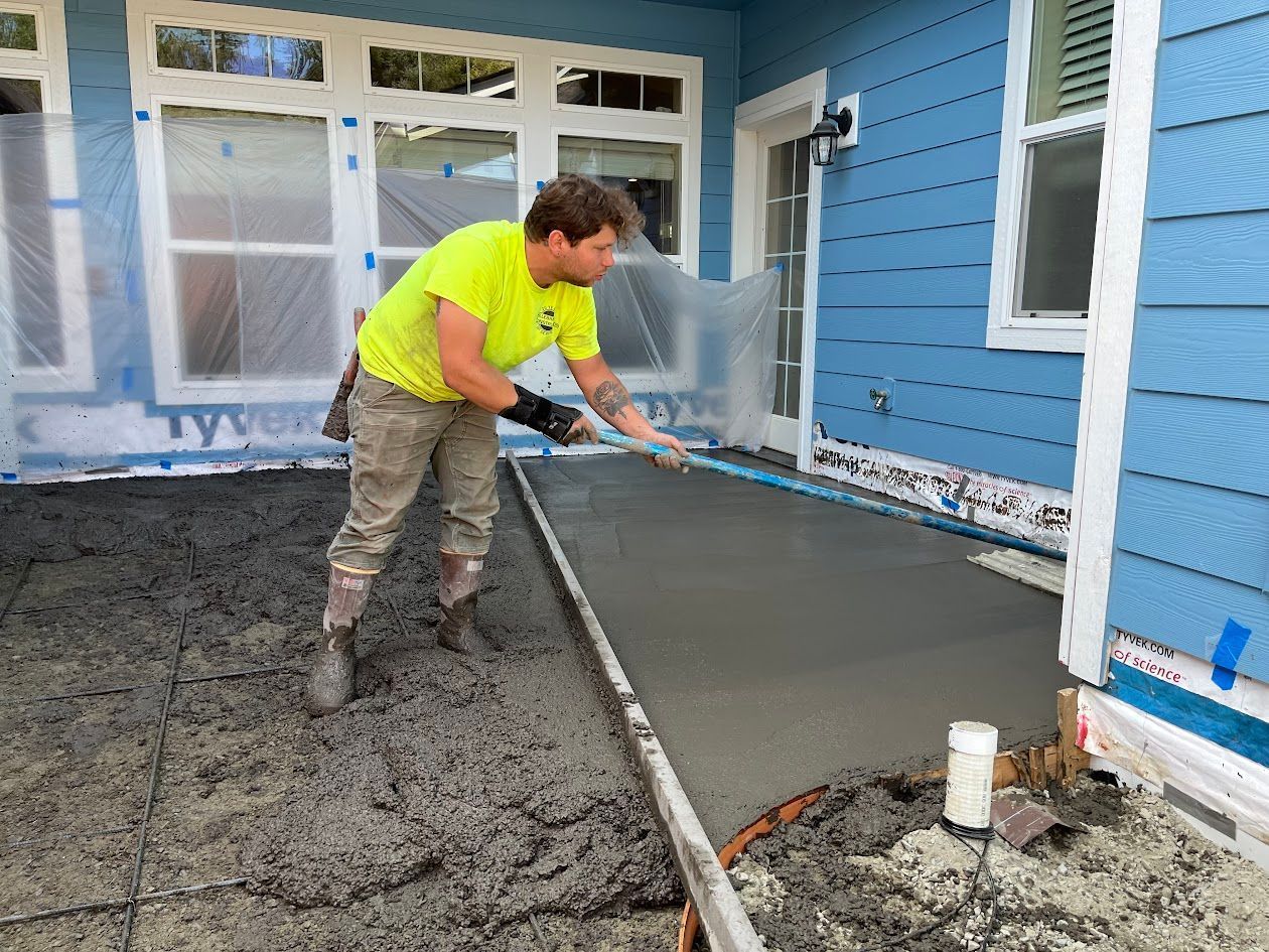 A man is spreading concrete on a sidewalk in front of a blue house.