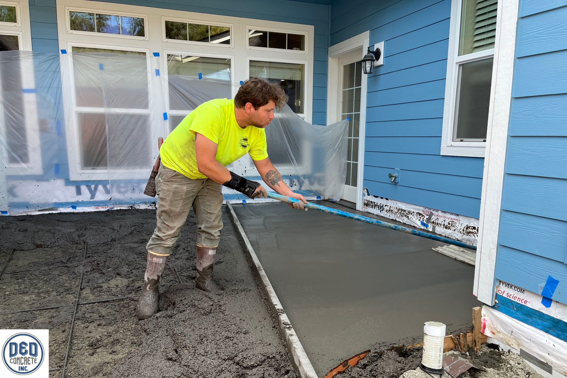 A man is pouring concrete on a sidewalk in front of a blue house.