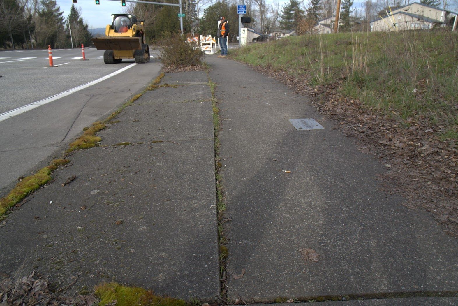A man is standing on a sidewalk next to a road while a bulldozer is working on the sidewalk.