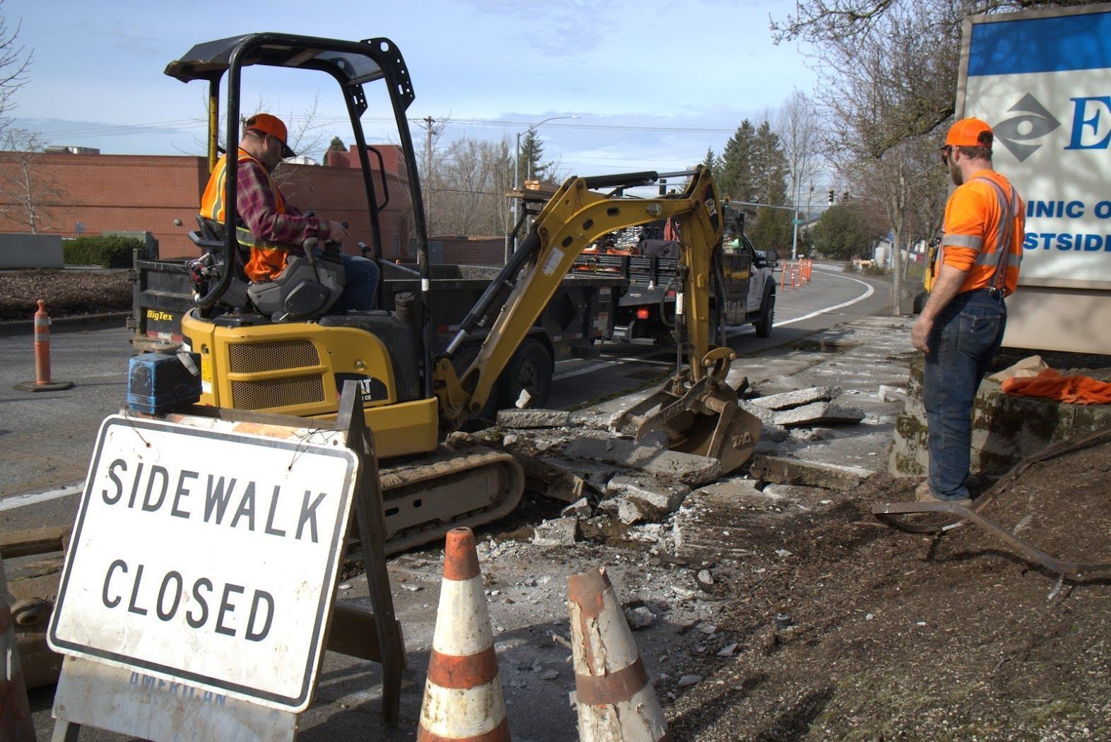 A man is driving a yellow excavator next to a sidewalk closed sign.