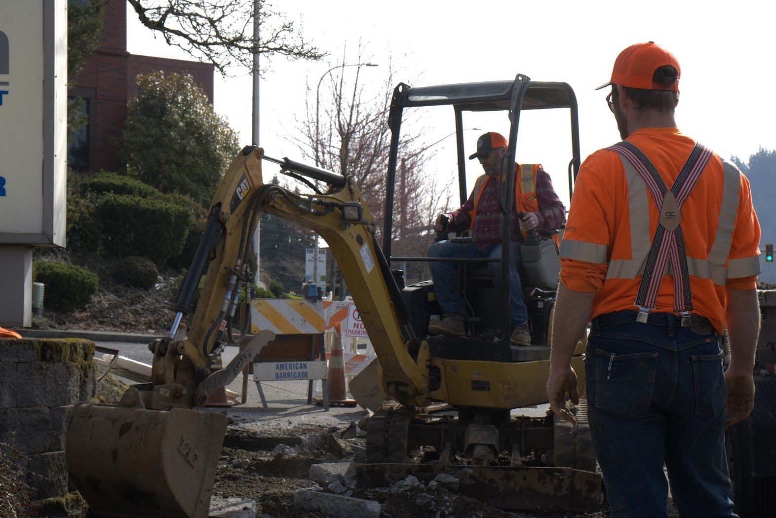 A man in an orange shirt is standing in front of an excavator