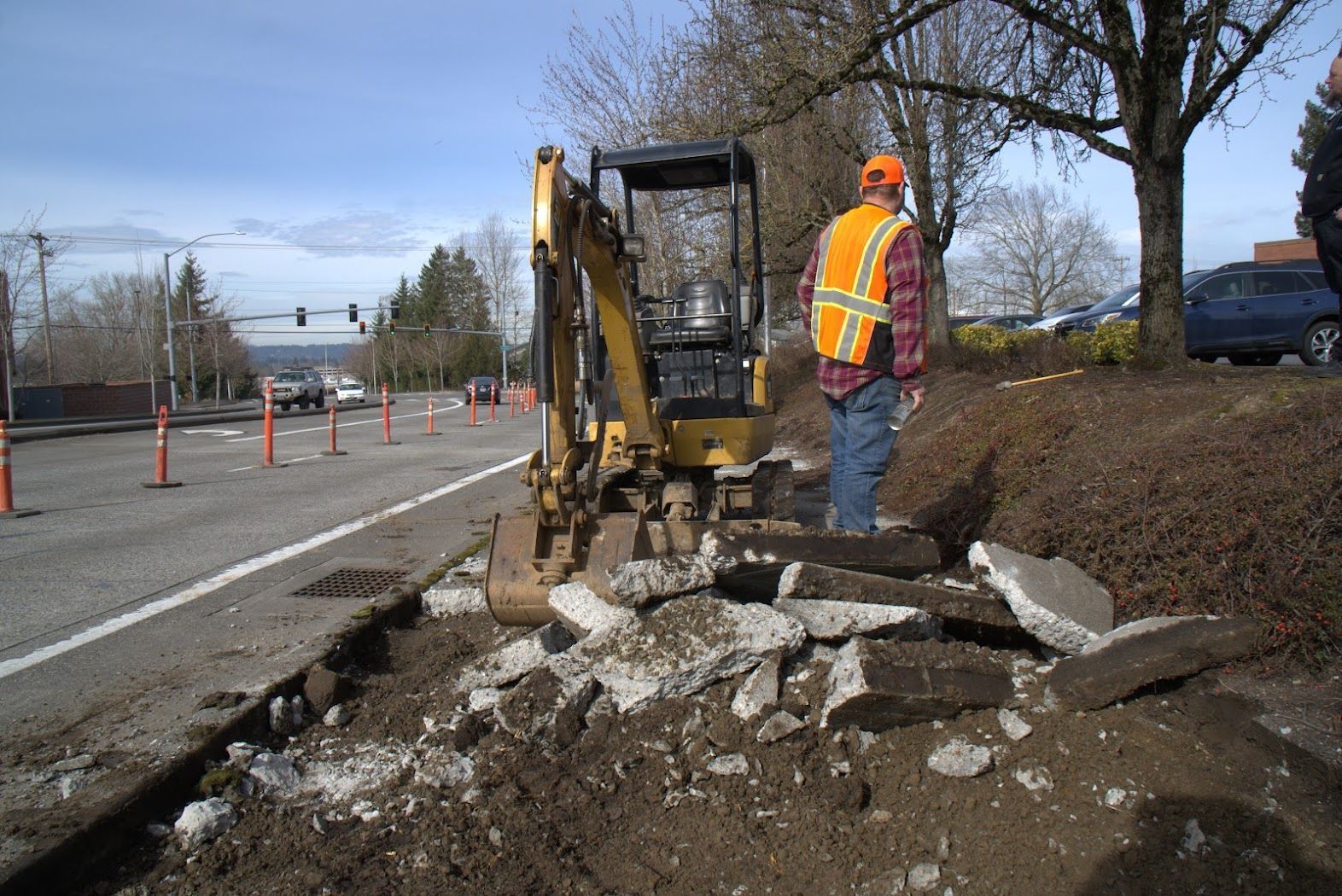 A man in an orange vest is standing next to a yellow excavator.
