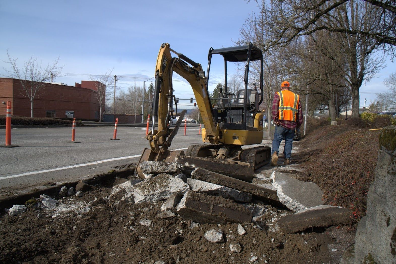 A man in an orange vest is standing next to a small excavator.