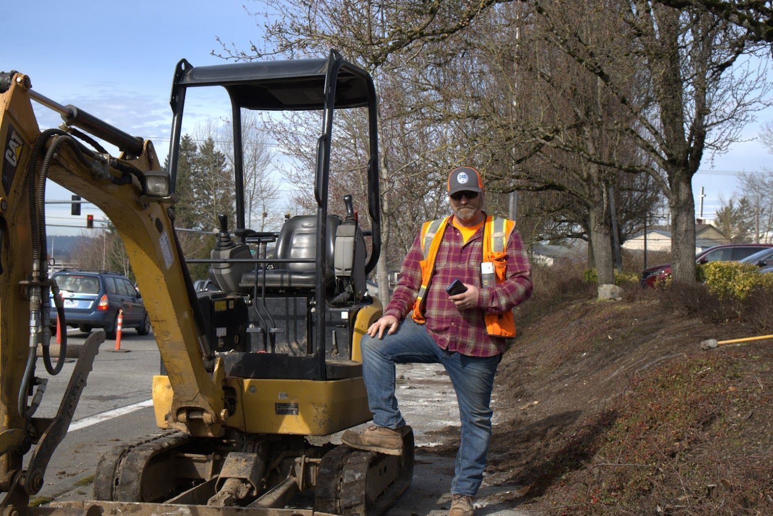 A man in an orange vest is standing next to a small excavator.