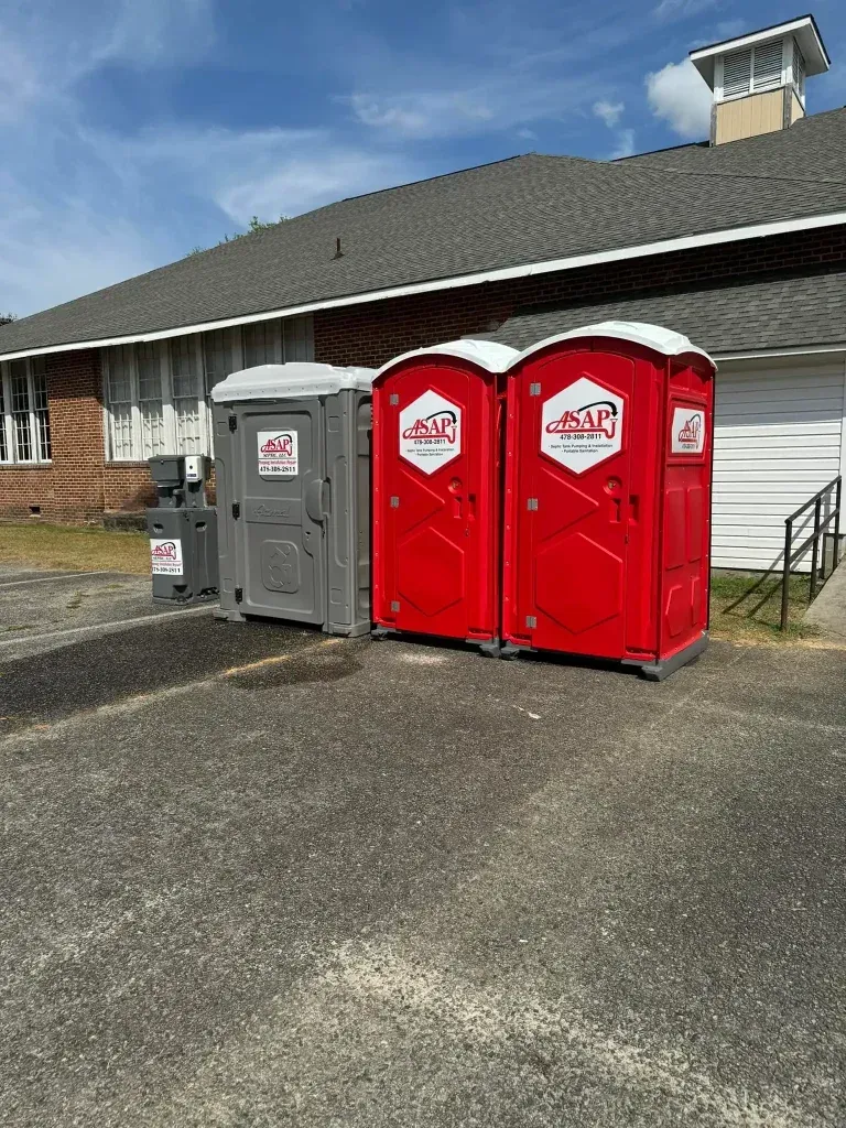 Three portable toilets, red and gray, parked outside a building on asphalt, under a blue sky.