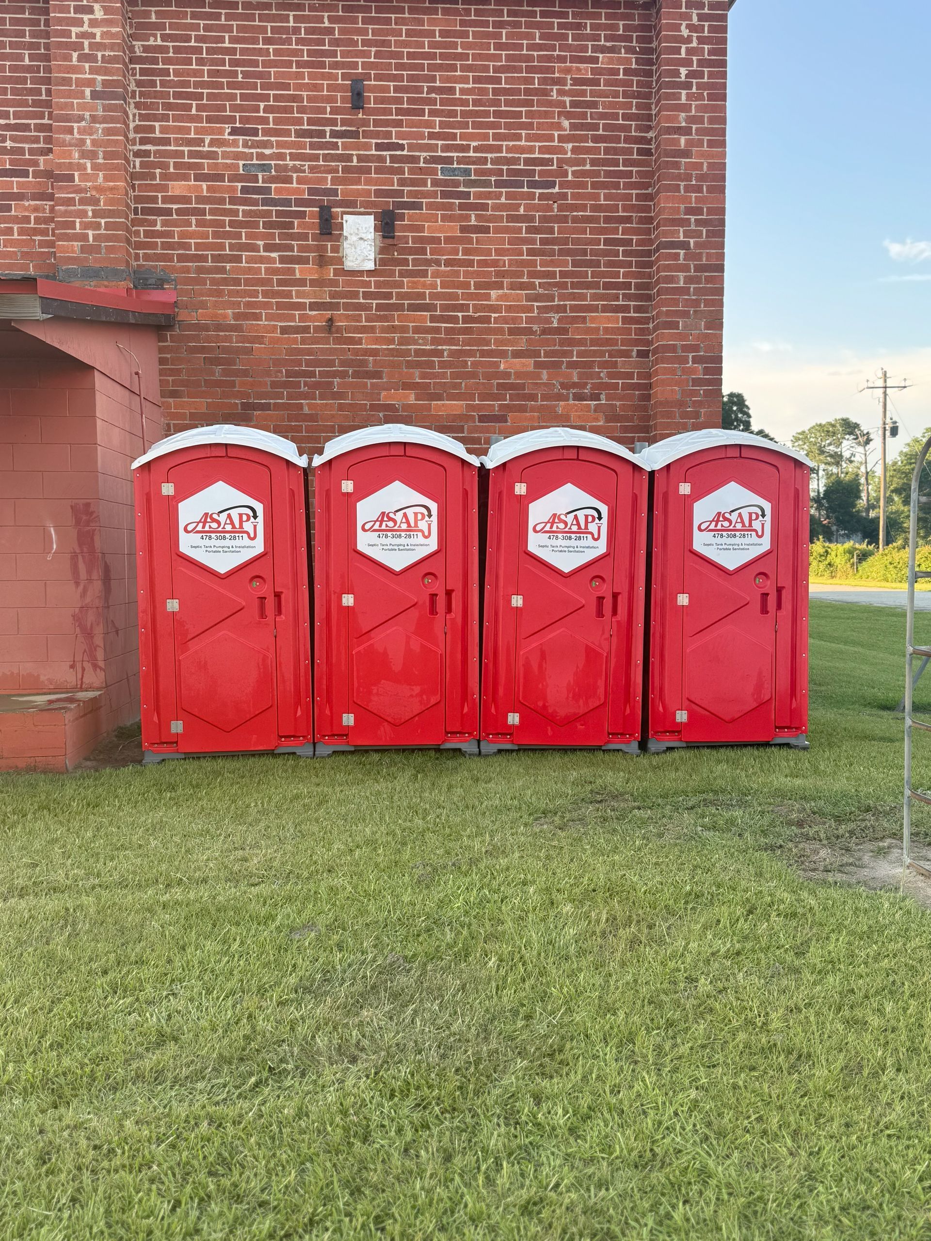 Four red portable toilets with logos are lined up on grass next to a brick building.