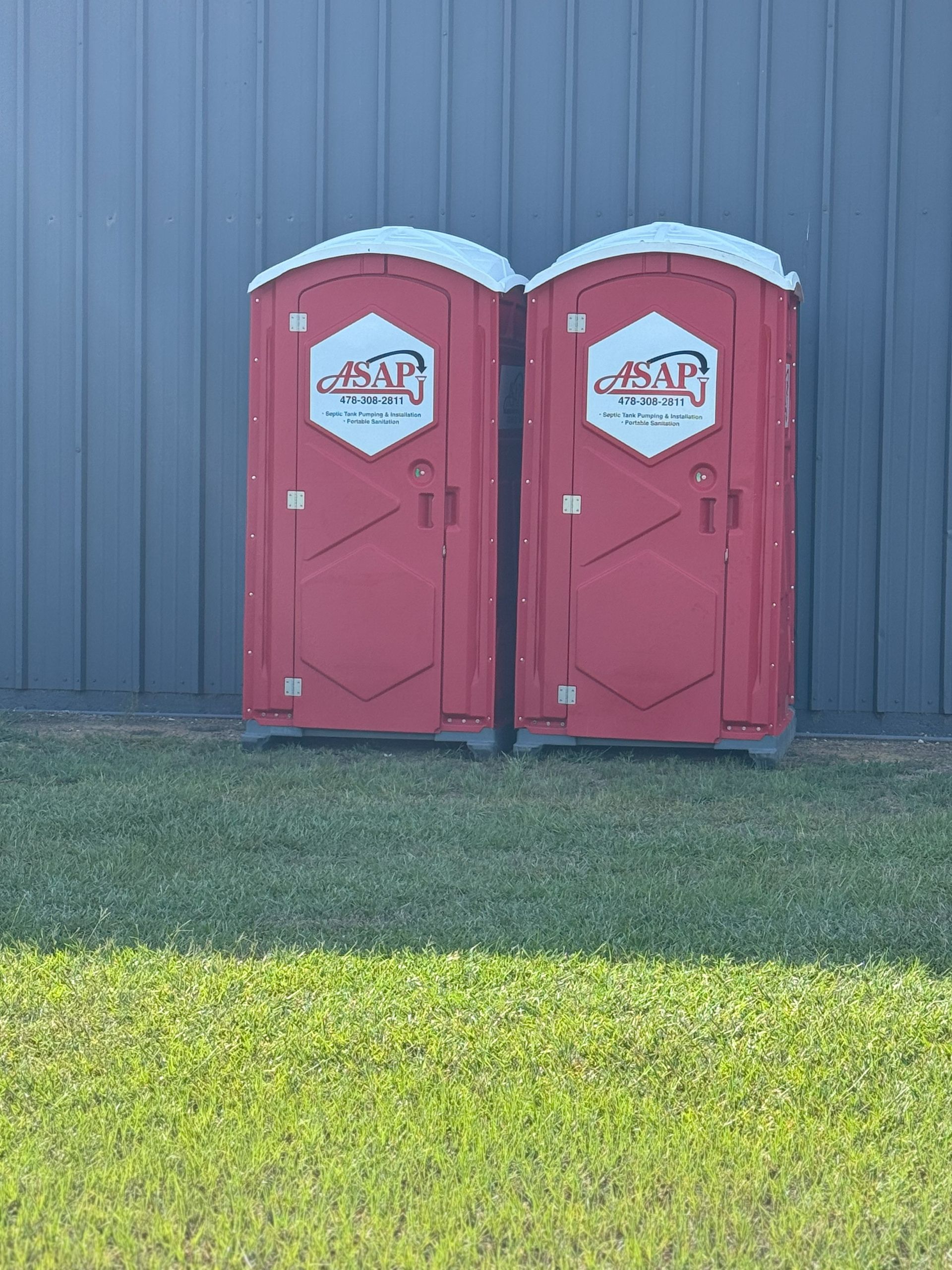 Two red portable toilets with logos on a grassy area, against a gray building.