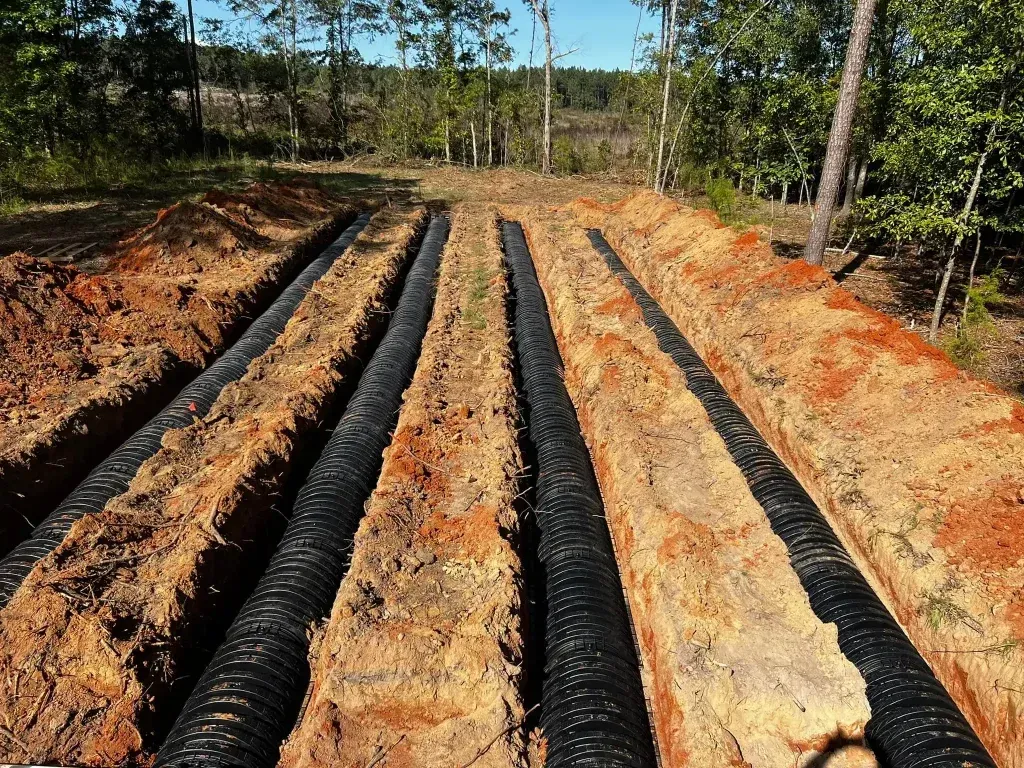 Four parallel trenches dug in earth with black corrugated drainage pipes laid within, in a wooded area.