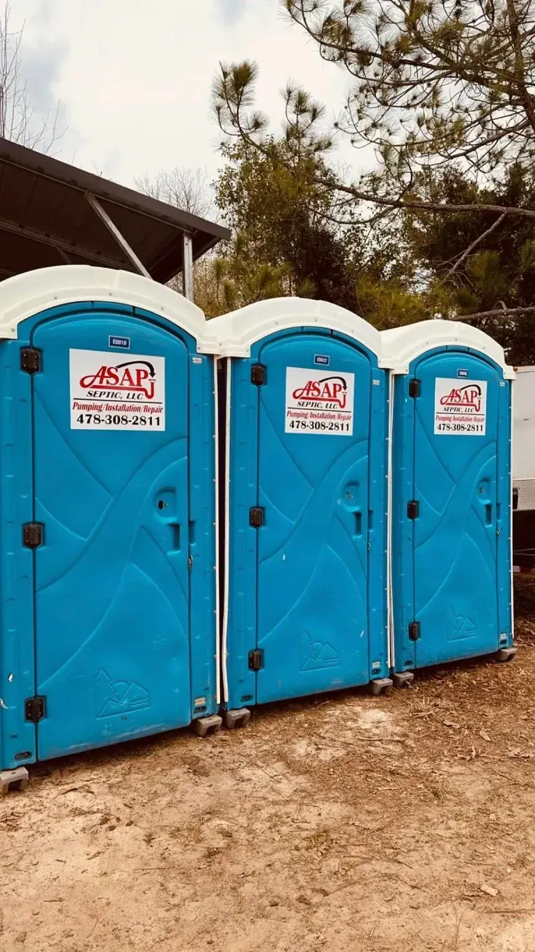 Three blue portable toilets in a row with company logos. Set outdoors, with trees in the background.
