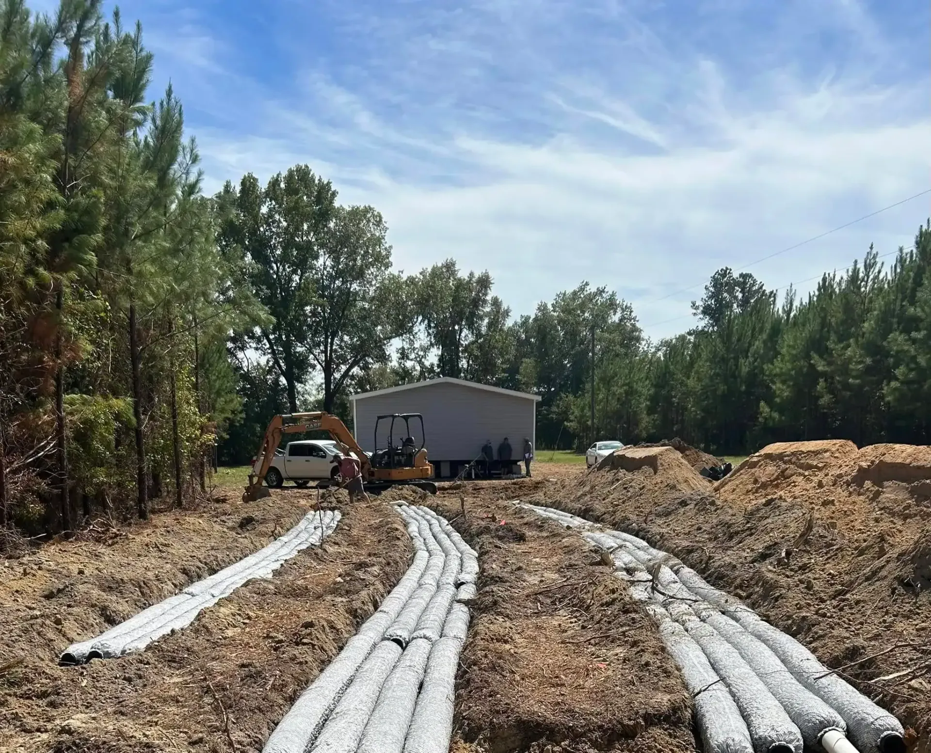 Construction site with a building and backhoe. Rows of gray drainage pipes lie on the ground, surrounded by mulch and trees under a blue sky.