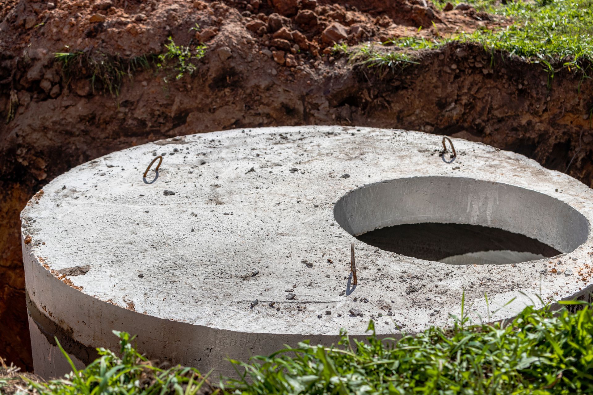 Person looking into an open septic tank. The tank opening is surrounded by grass and leaves.