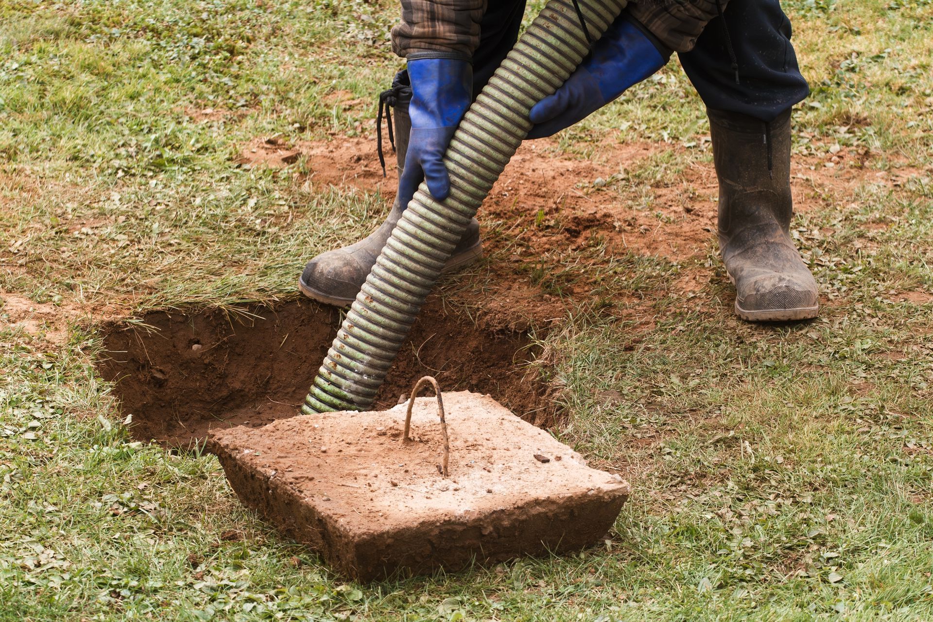 Person in boots and gloves pumping septic tank, lid and hole in grass.