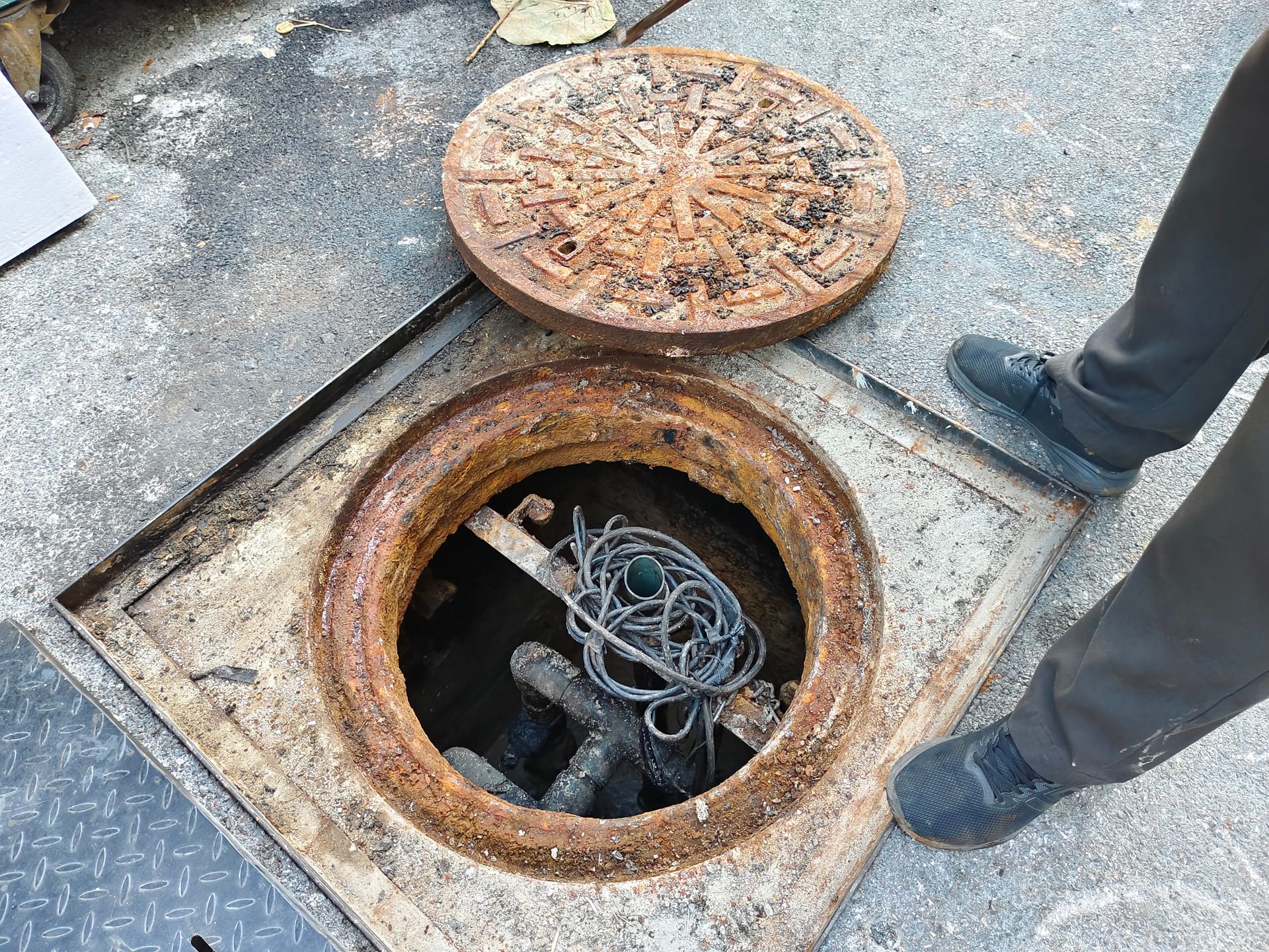 Open sewer manhole with rusty cover. A person stands nearby. Cables are inside.