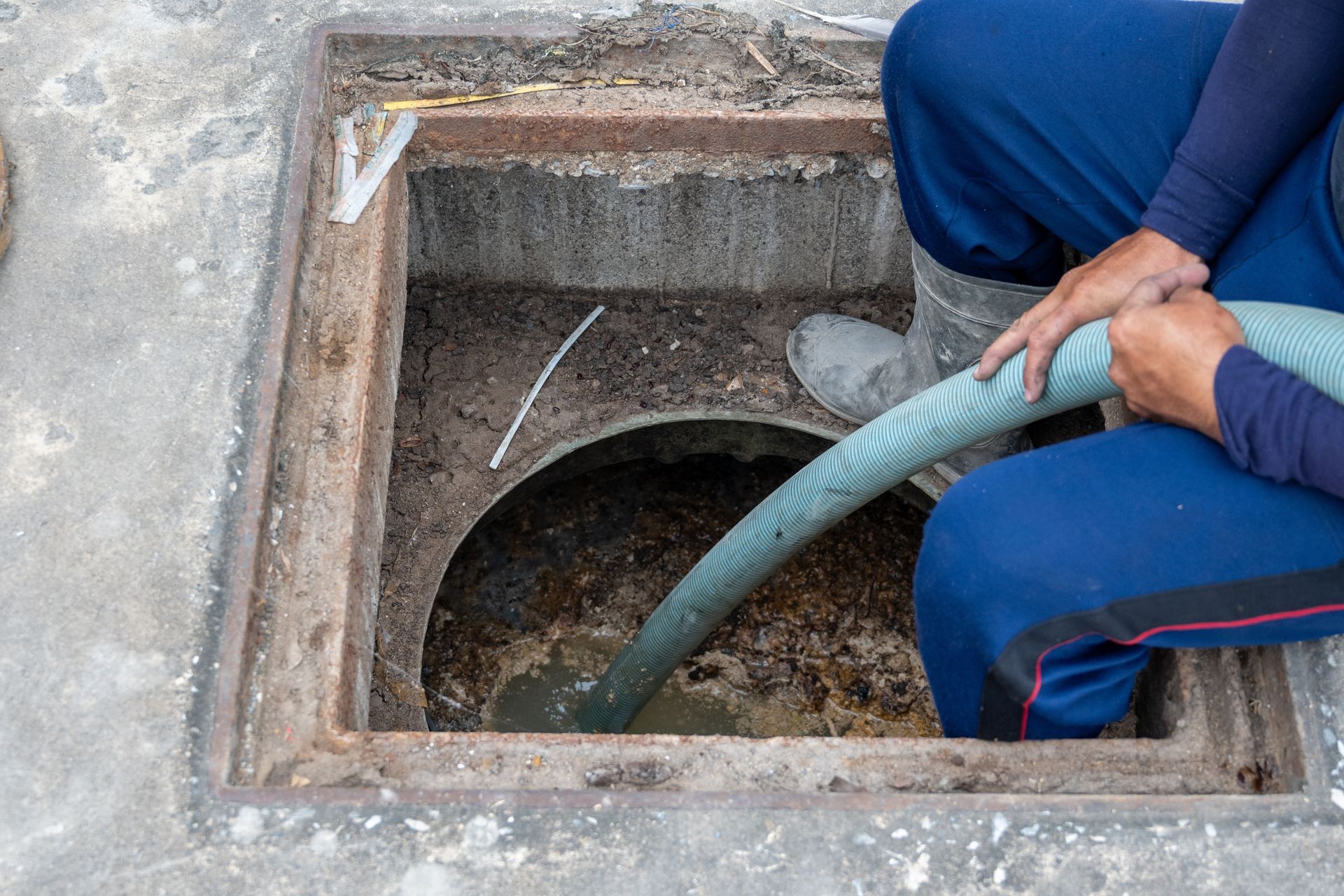 Person pumping sewage from an open manhole. The manhole is square-shaped, surrounded by concrete.