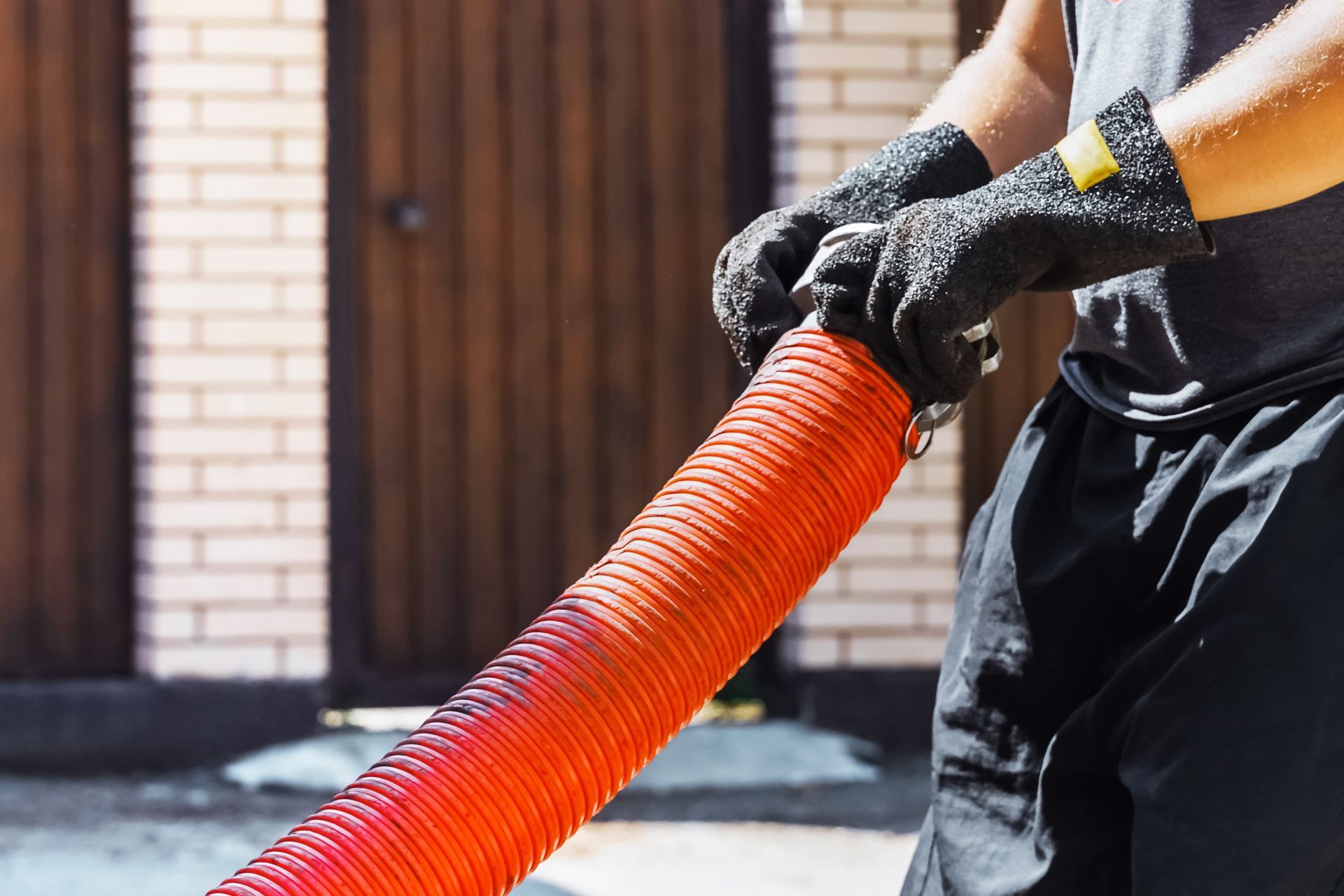 Person in gloves holding a large, red corrugated hose near a brick building.