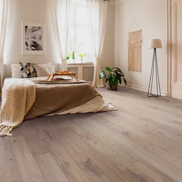 Bedroom with light wood floors, bed with beige bedding, large window with curtains, neutral decor.