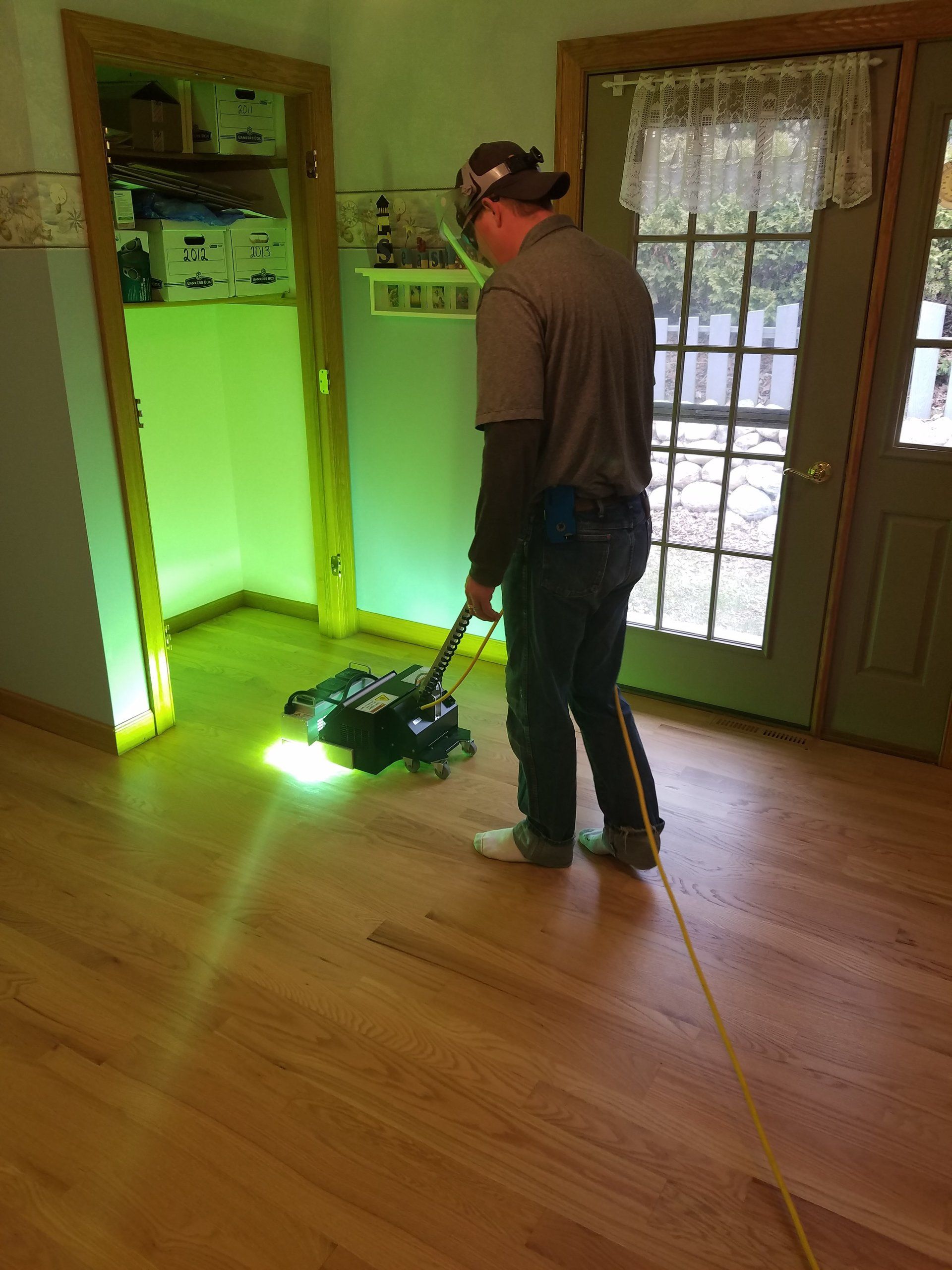 A man using a floor polisher on hardwood in a room with a green-lit doorway and a wooden door.