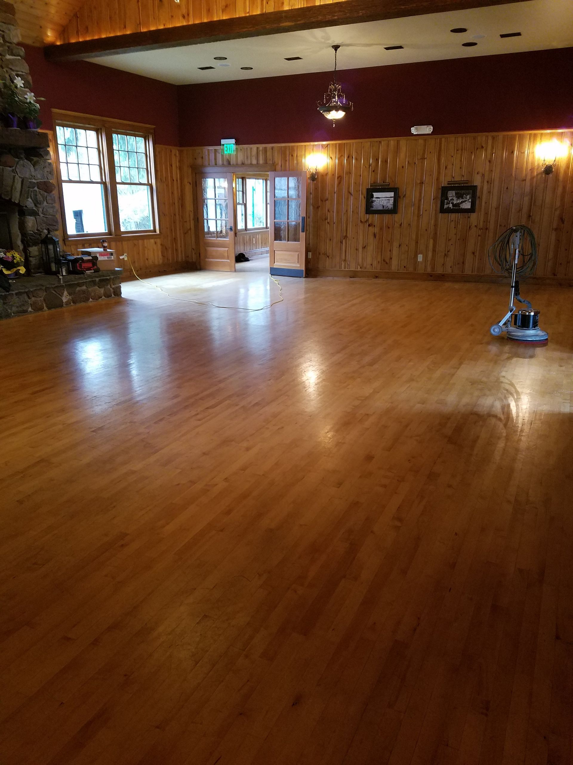 Spacious room with wooden floor and walls. Large stone fireplace, windows, and a door.