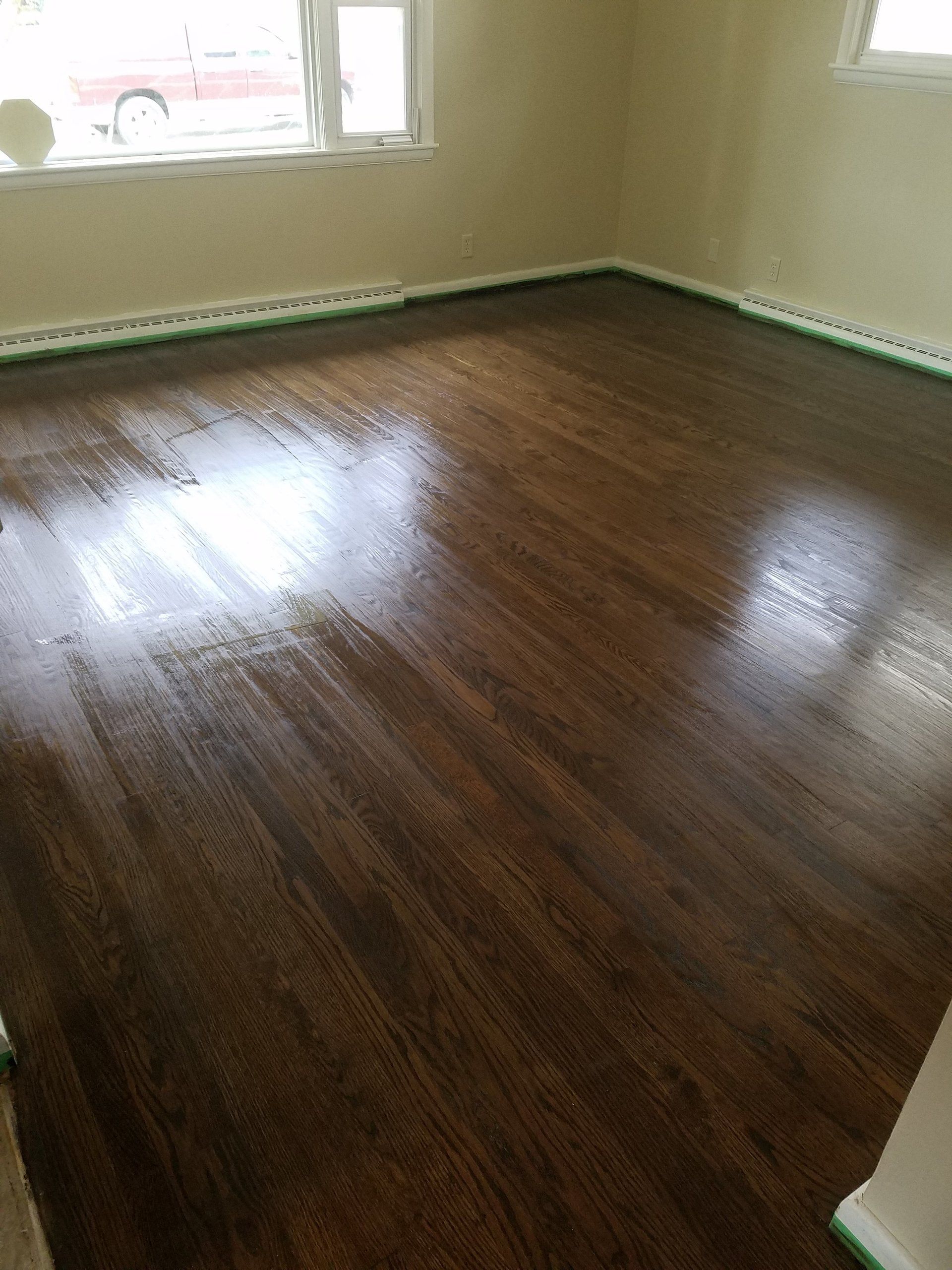 Dark stained hardwood floor in a room, reflecting light. Green tape along the baseboards.