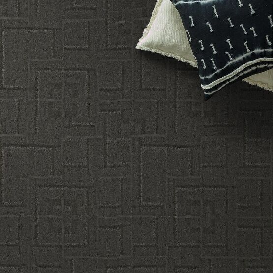 Dark gray geometric patterned carpet with a patterned pillow on top.