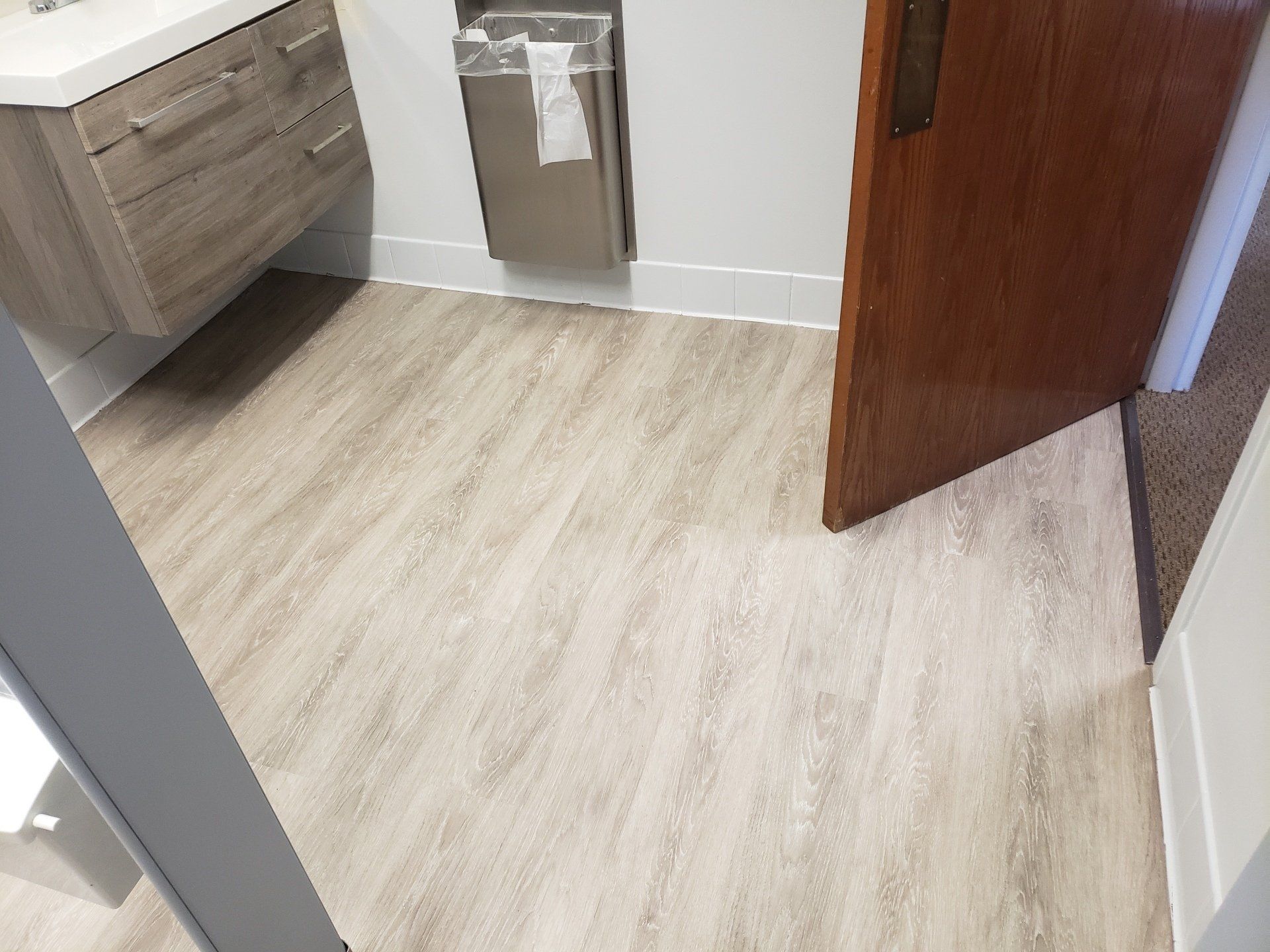 Bathroom with wood-look flooring, vanity, brown door, and stainless steel trash can.
