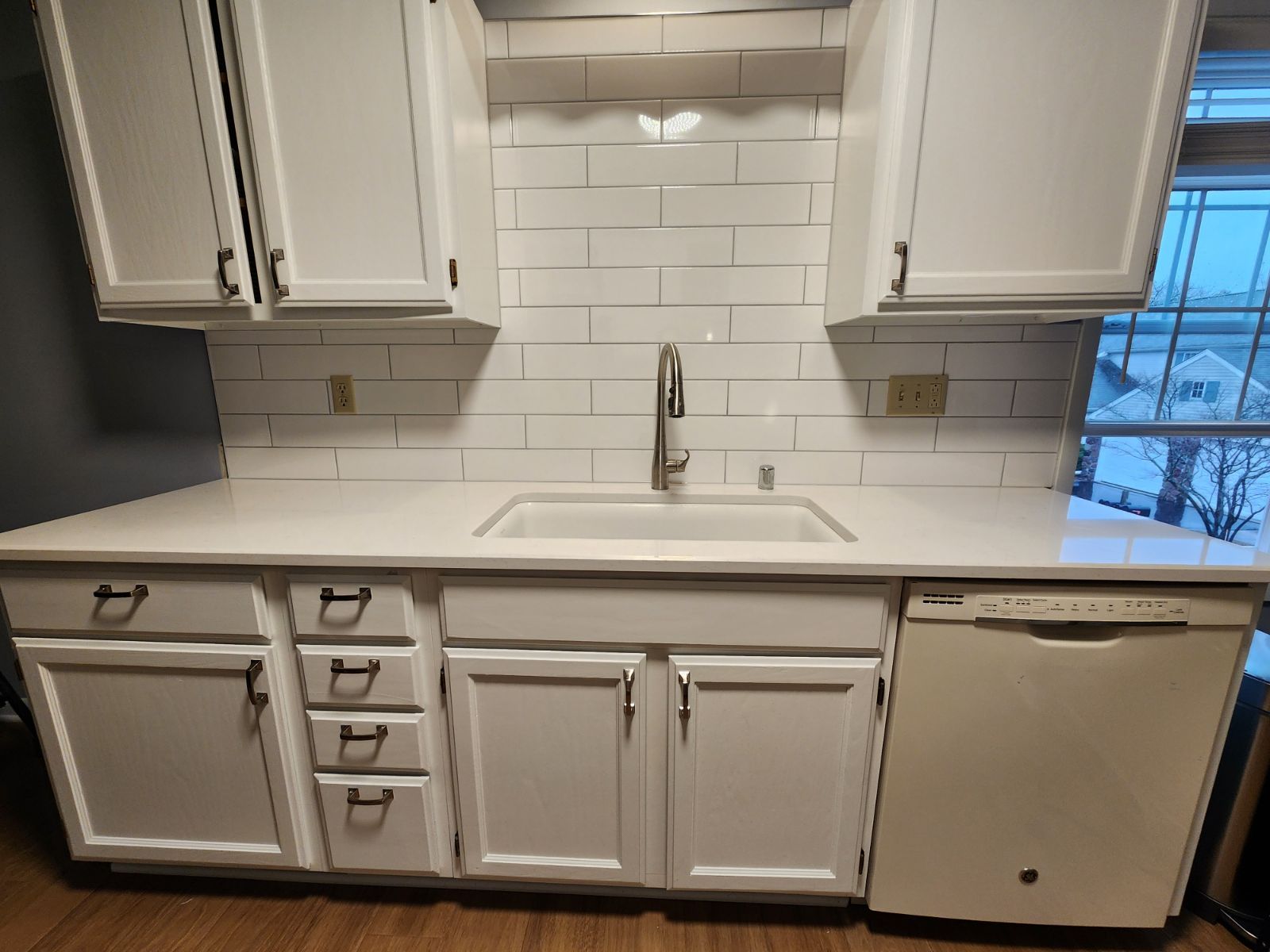 White kitchen cabinets and sink with white subway tile backsplash and a window to the right.