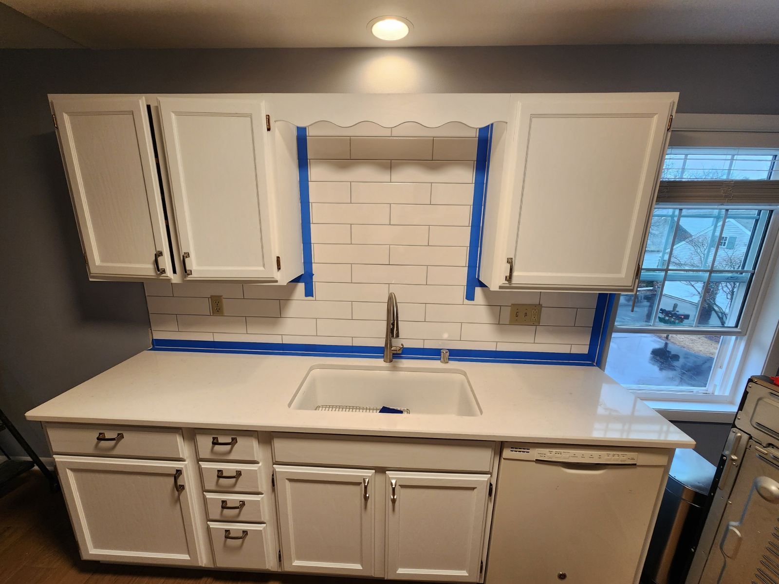 White kitchen cabinets and countertop, blue tape, subway tile backsplash, stainless steel sink and faucet.