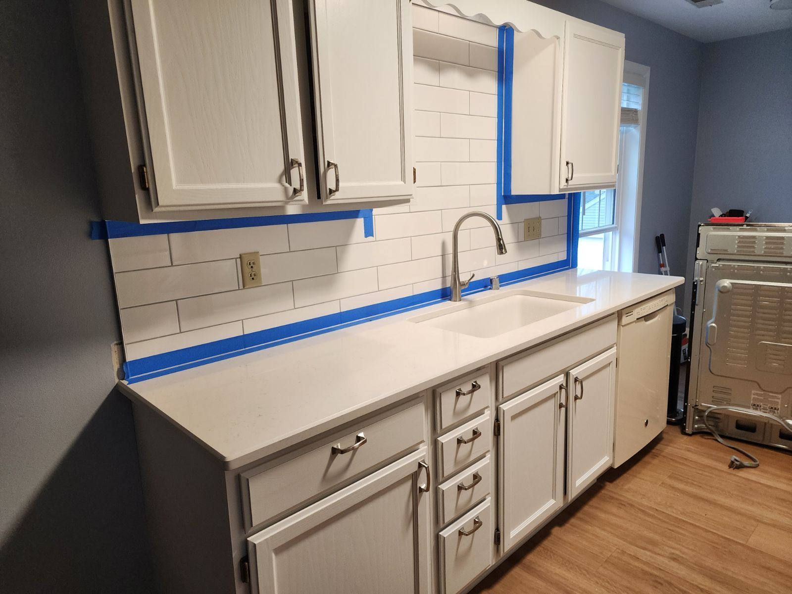 Kitchen renovation: white cabinets, countertops, and backsplash tiles, with blue painter's tape along edges.