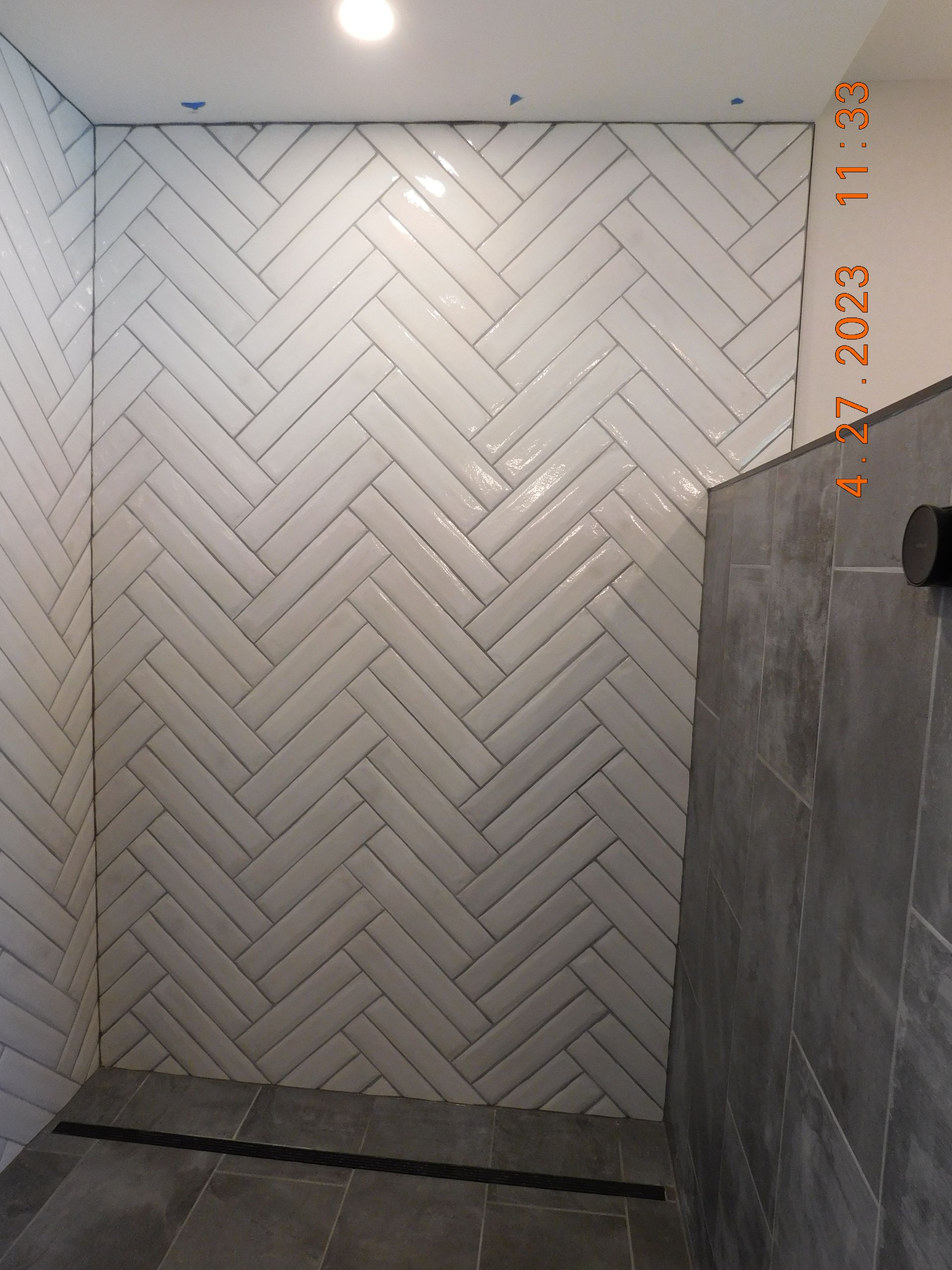 Herringbone-tiled shower wall; white tiles, gray floor and wall, recessed lighting, and black drain.