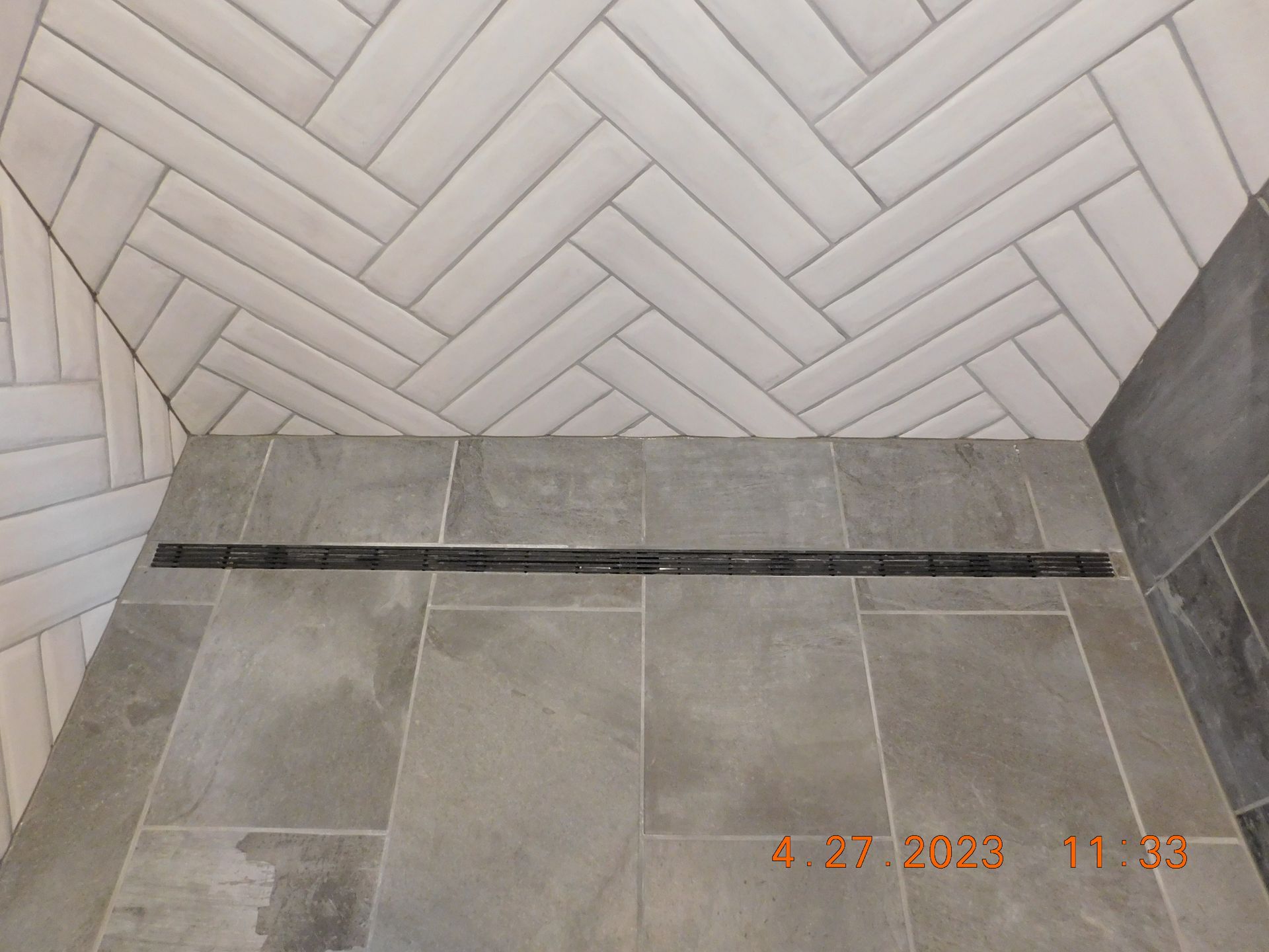 Shower with gray tiled floor and herringbone-patterned white wall tiles, a linear drain, and dark gray side tiles.