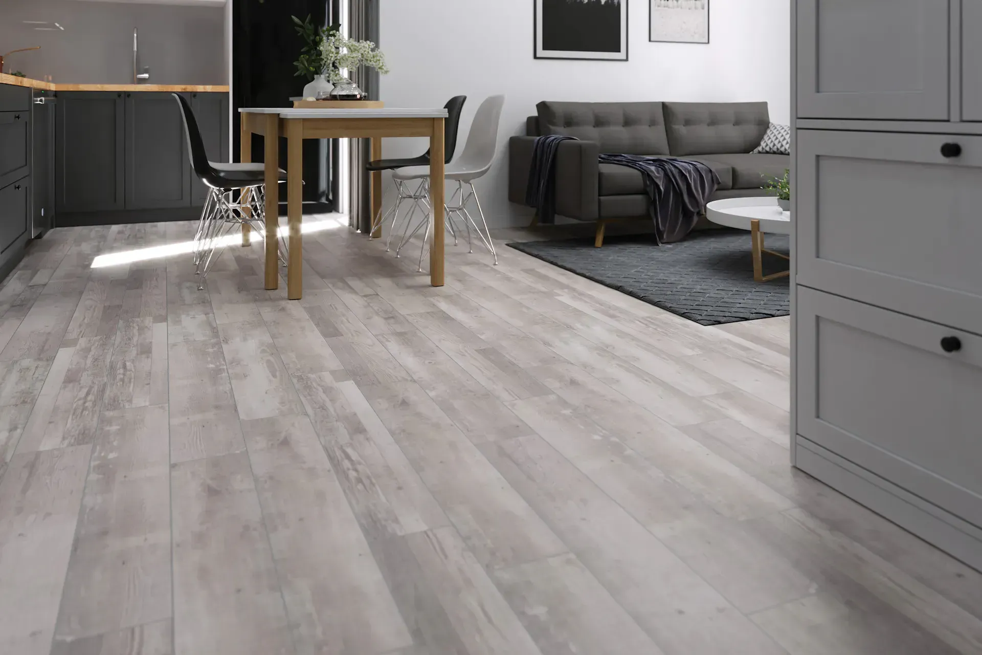 Light-colored wood flooring in a modern living space with a dining area, gray sofa, and kitchen cabinets.