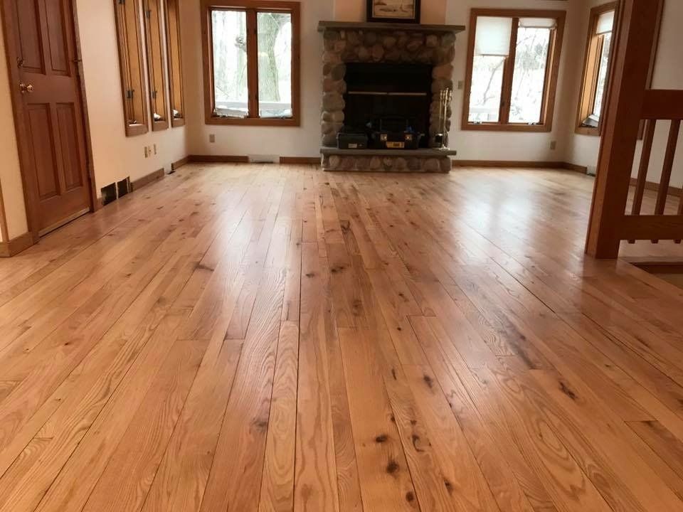 Light wood floor in a living room with a stone fireplace, windows, and a wooden door.
