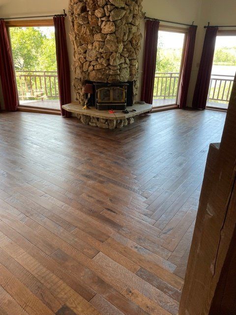 Herringbone wood floor in room with stone fireplace and large windows overlooking a deck.