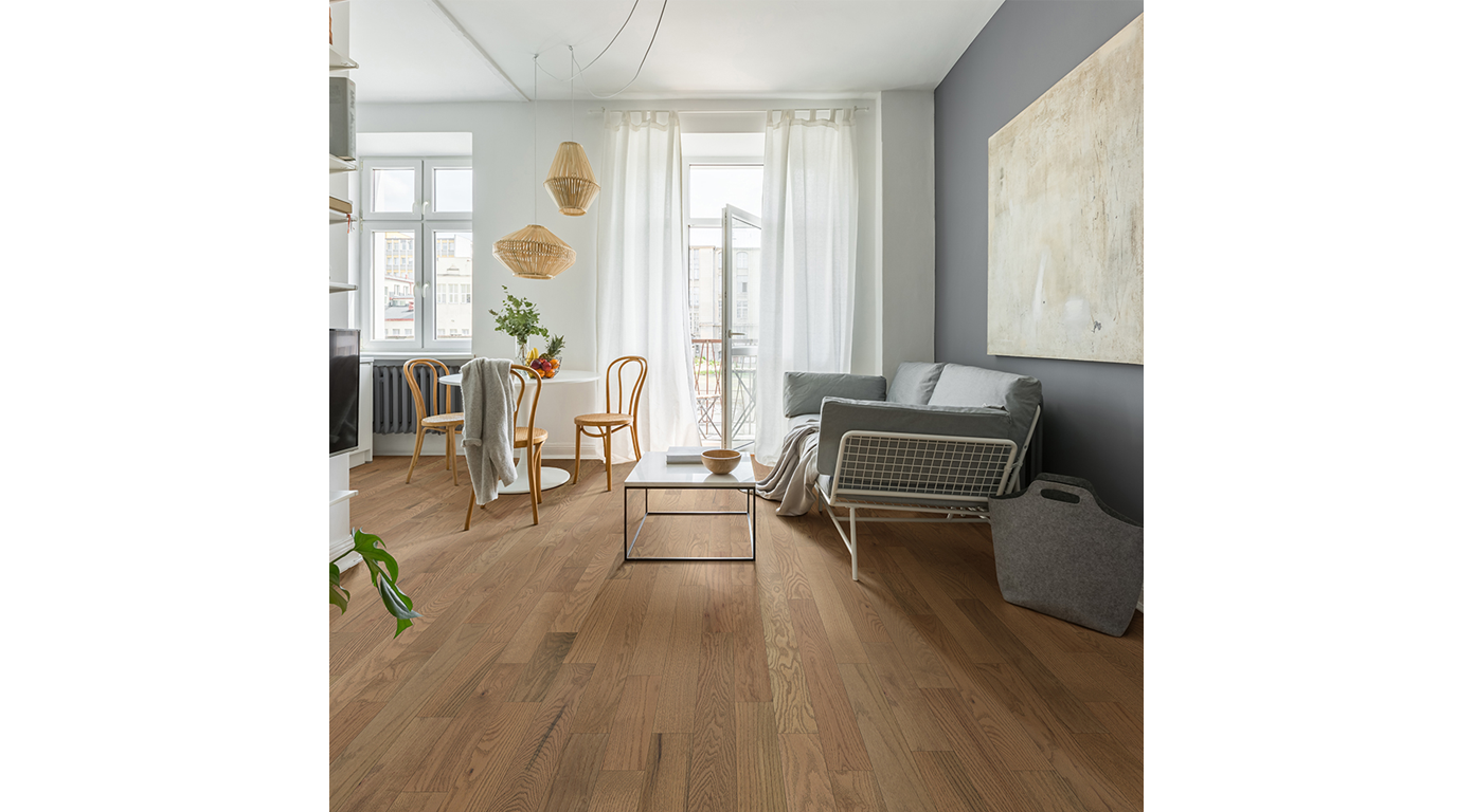 Living room with wooden floors, a gray couch, and neutral-toned decor. Sunlight streams through the windows.