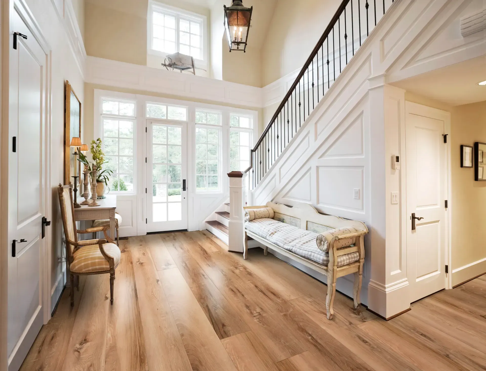 A bright entryway with light wood floors, white walls, and a staircase. A bench and chair sit along the walls.