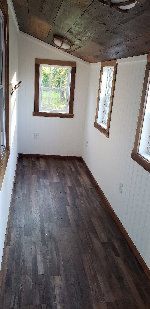 An interior shot of a small room with wood flooring, white walls, and wood-framed windows. The ceiling is wood.
